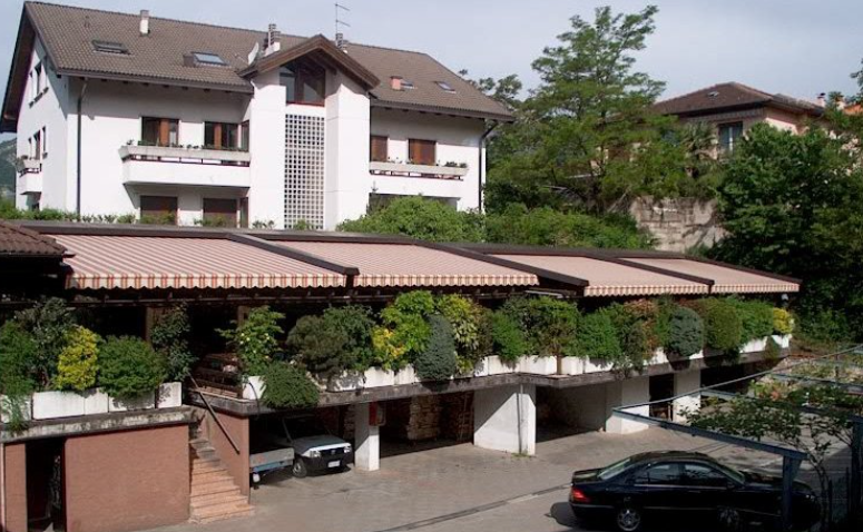 White building with balconies, awnings, and plants, with a car in the foreground.