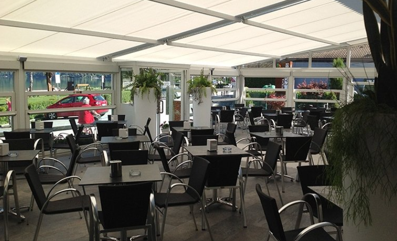 Outdoor cafe with tables and chairs under a white canopy. Sunlight streams in.