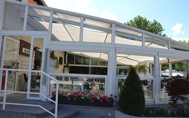 Glass-enclosed cafe with white frames, awnings, and plants outside. Entrance has a white ramp and door.