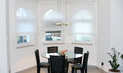 Dining area with round glass table, black chairs, white arched windows with blinds, and a modern light fixture.
