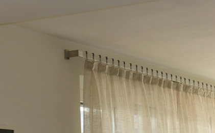 Beige sheer curtains hanging from a silver rod, mounted on a white wall near a window.