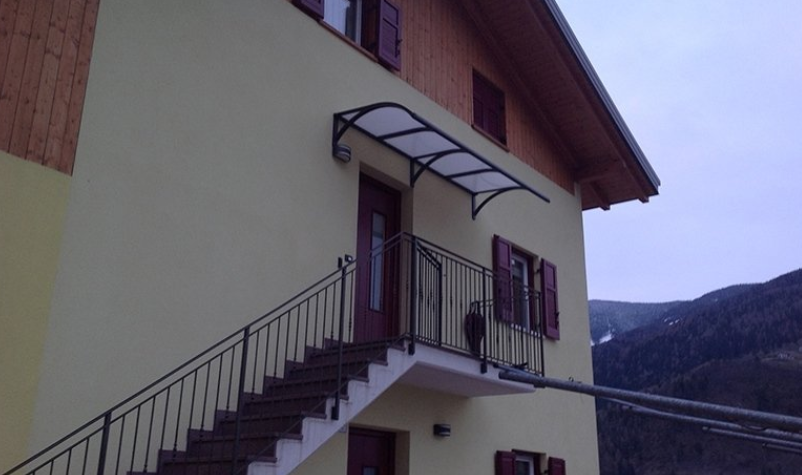 Yellow building with stairs leading to a door under a clear awning. Black railing along stairs and balcony.