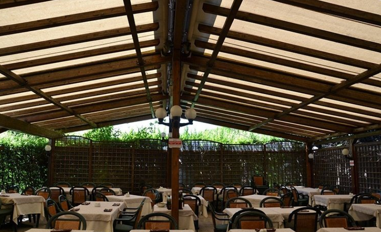 Outdoor restaurant with tables covered in white tablecloths beneath a wooden canopy.