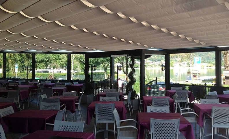 Restaurant patio with tables set for service, overlooking a lake. Beige retractable awning, white chairs, red tablecloths.