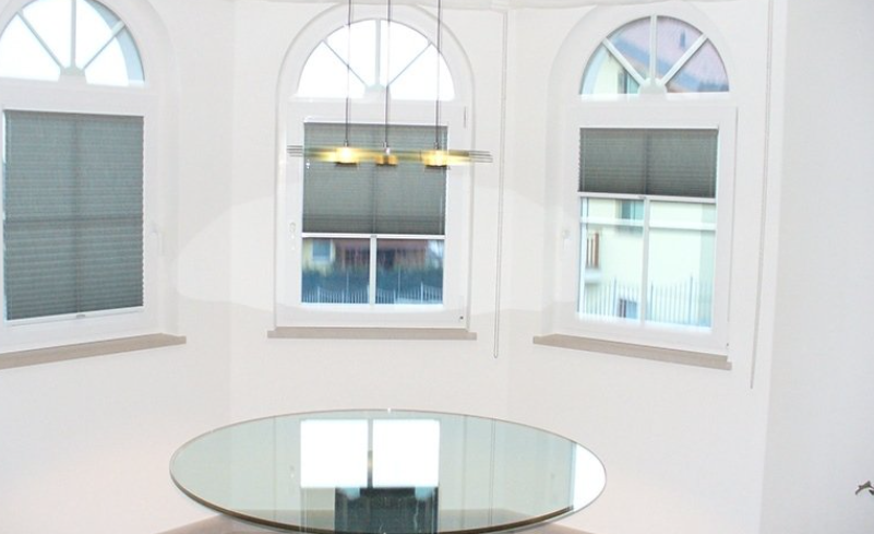 A room with three arched windows, gray shades, and a large round glass table.
