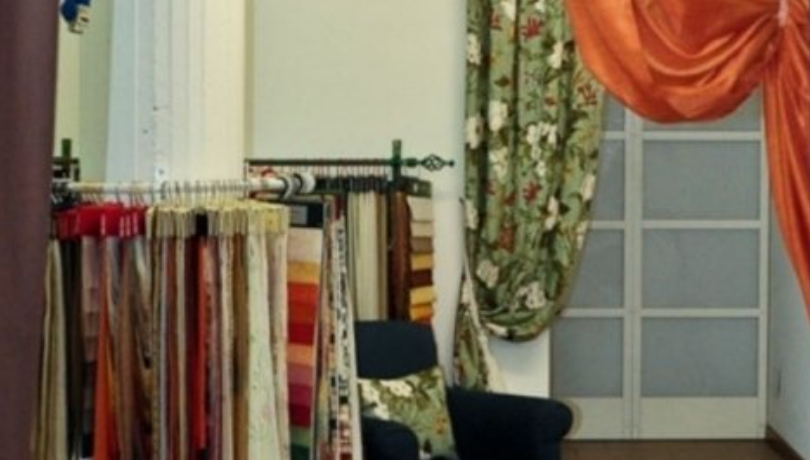 Fabric samples hang on a rack in a showroom, alongside draped curtains and a chair.