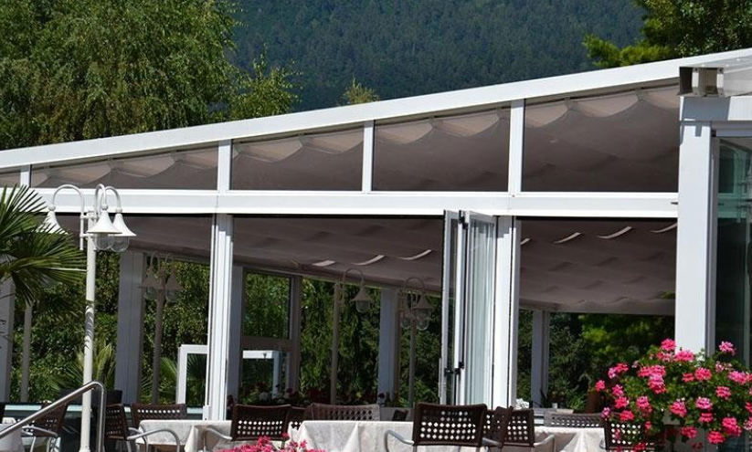 White-framed outdoor dining area with beige fabric covering. Tables are set, flowers and trees are in the background.