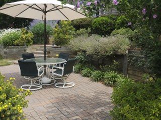 Patio with table, chairs, umbrella, surrounded by garden beds, brick patio, sunny.
