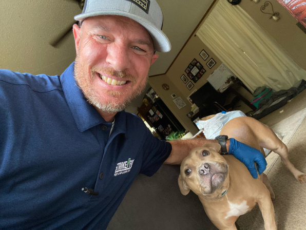 Man in blue shirt and cap smiles, arm around a tan dog in a home.