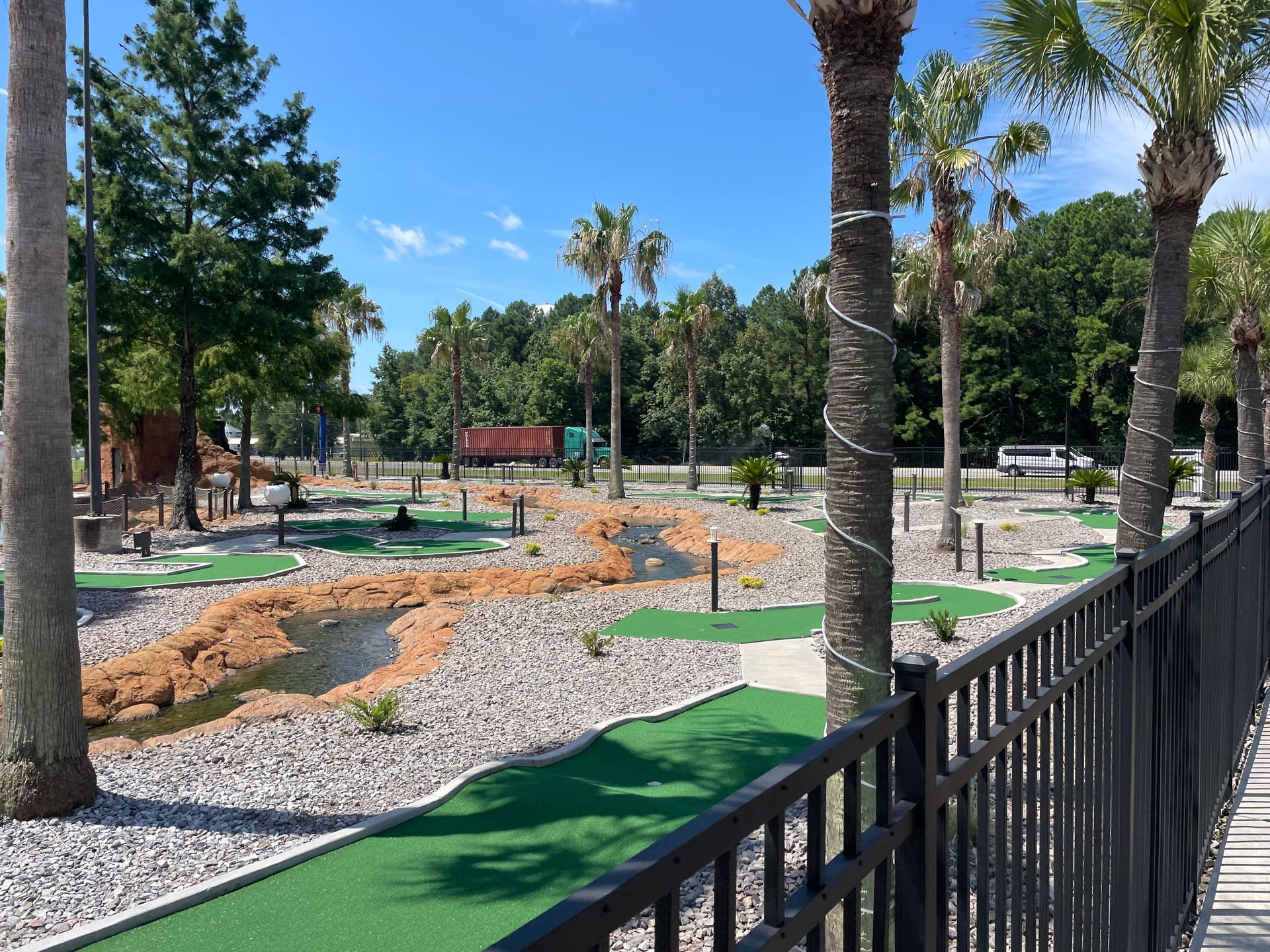 A fence surrounds a mini golf course with palm trees in the background.