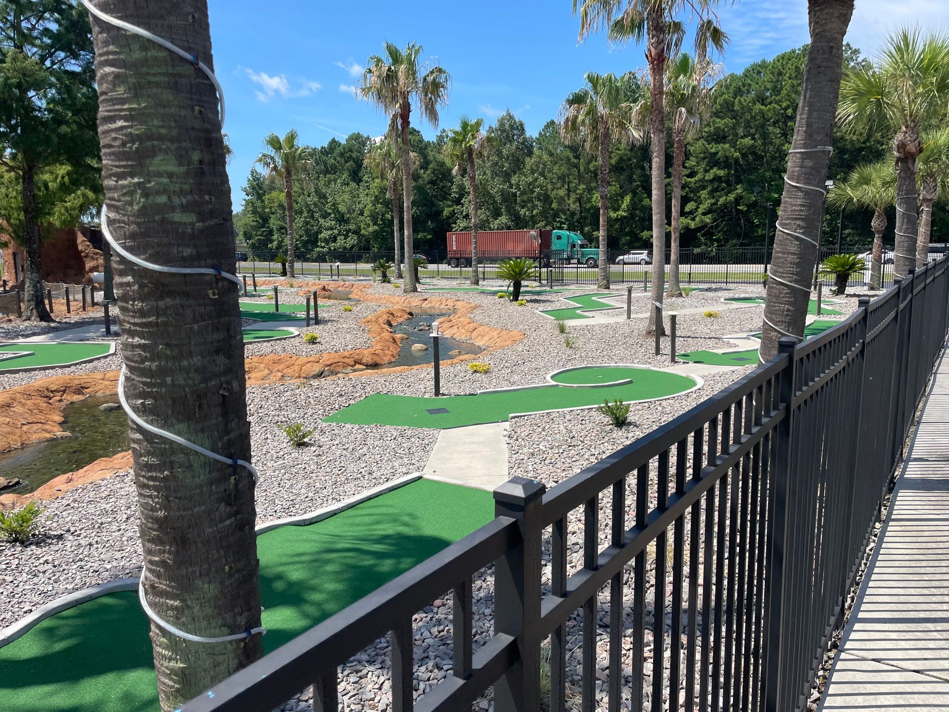A fence surrounds a mini golf course with palm trees in the background.