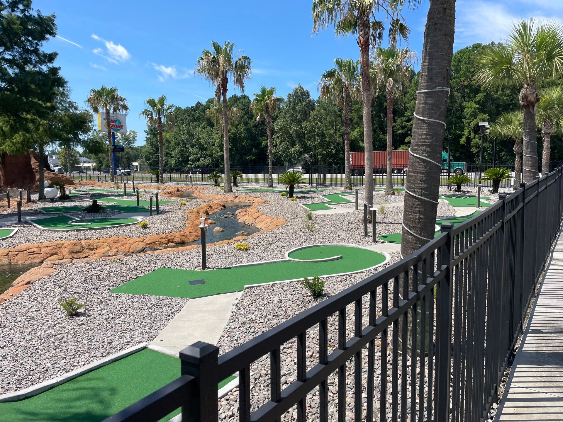 A fence surrounds a mini golf course with palm trees in the background.