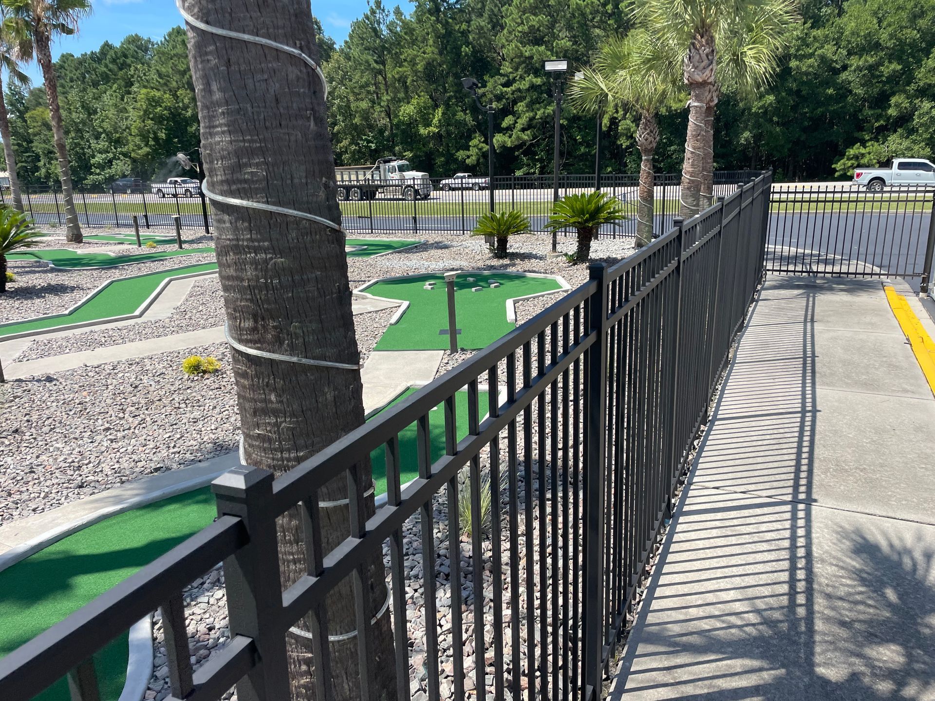 A metal fence surrounds a mini golf course with palm trees in the background.