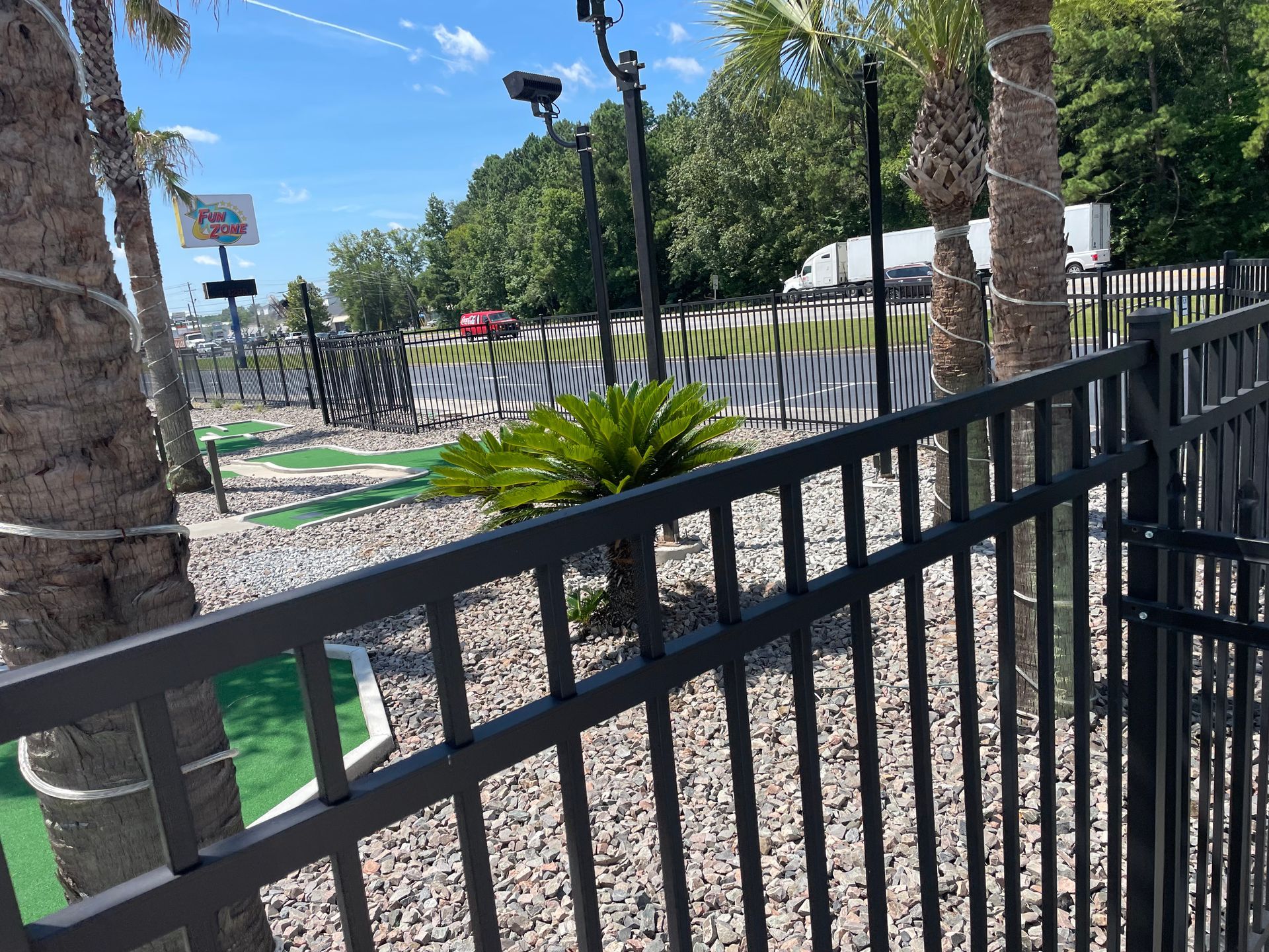 A black fence surrounds a mini golf course with palm trees in the background.