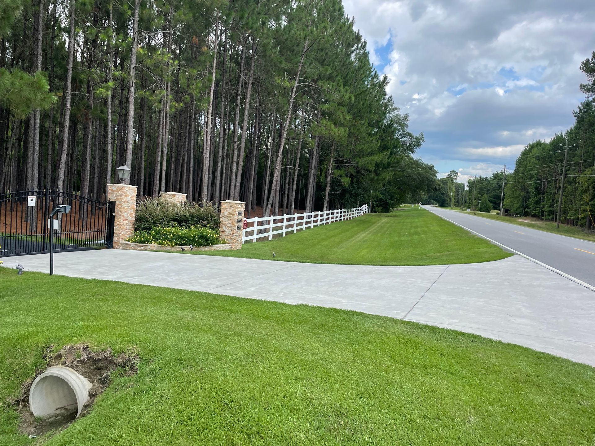 A concrete driveway leading to a fenced in area with trees in the background.