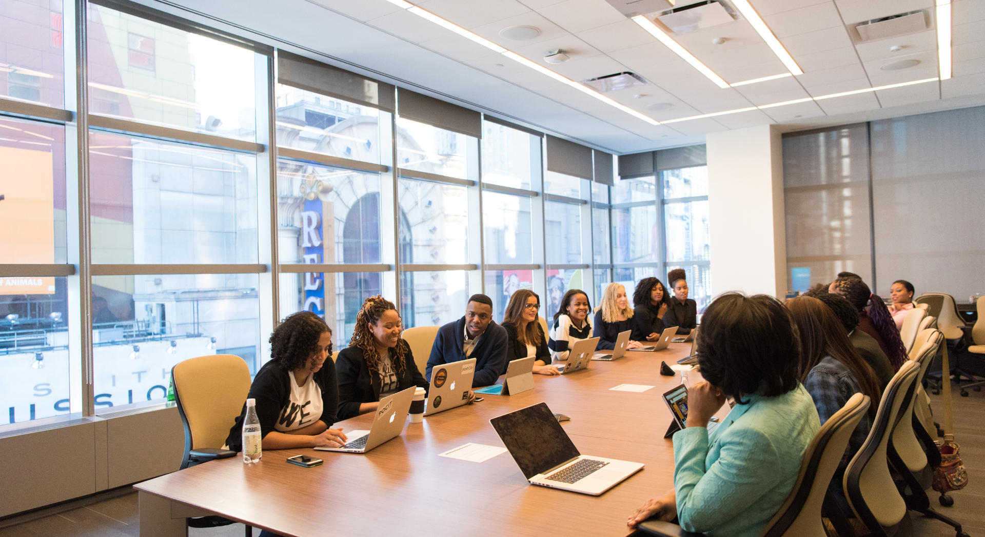A group of people are sitting at a long table in a conference room with laptops.