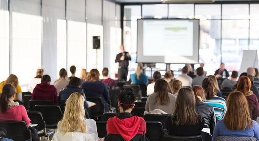 A group of people are sitting in a lecture hall watching a presentation.