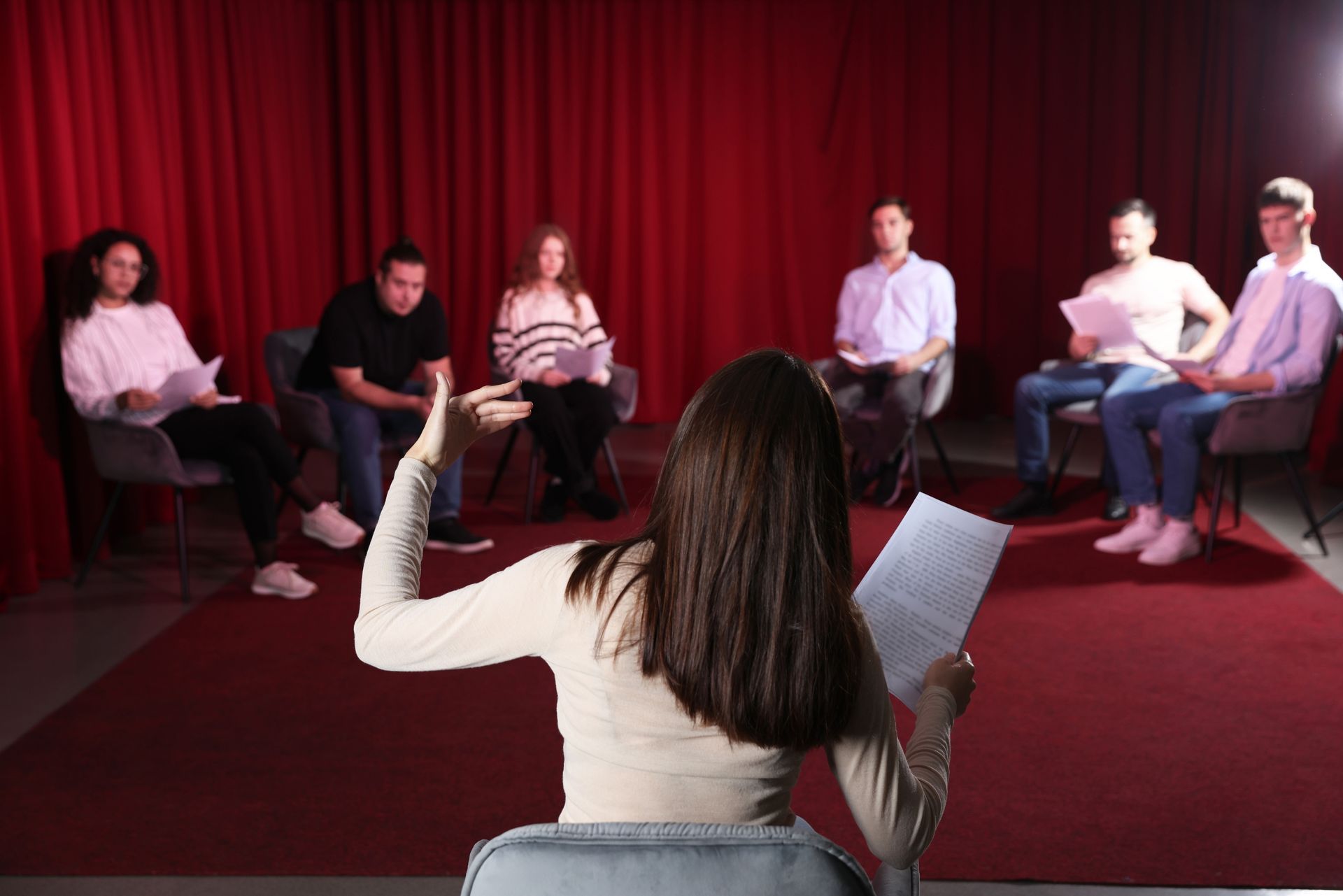 A woman is reading a script to a group of people sitting in chairs in front of a red curtain.