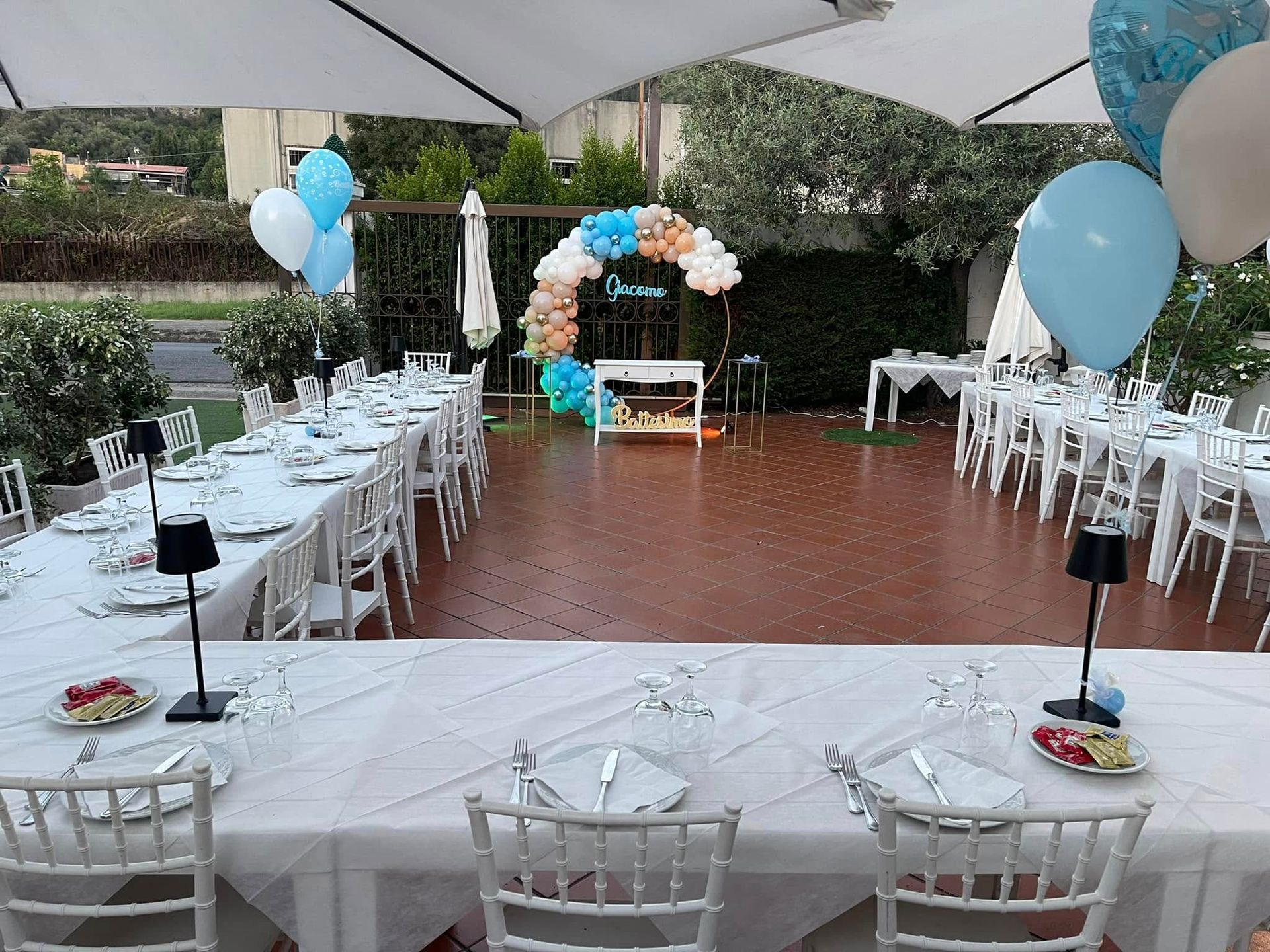 Tavoli per feste all'aperto apparecchiati con tovaglie bianche, sedie e decorazioni, tra cui palloncini e un arco.
