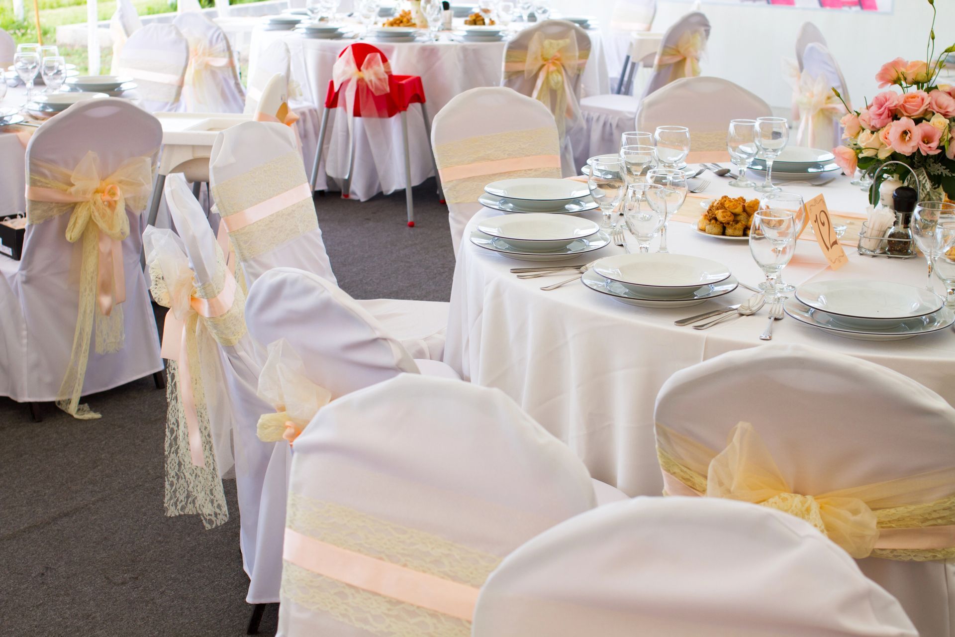 Tavoli e sedie con tovaglie bianche, decorati con nastri dai colori chiari, apparecchiati per un evento formale, con fiori al centro.