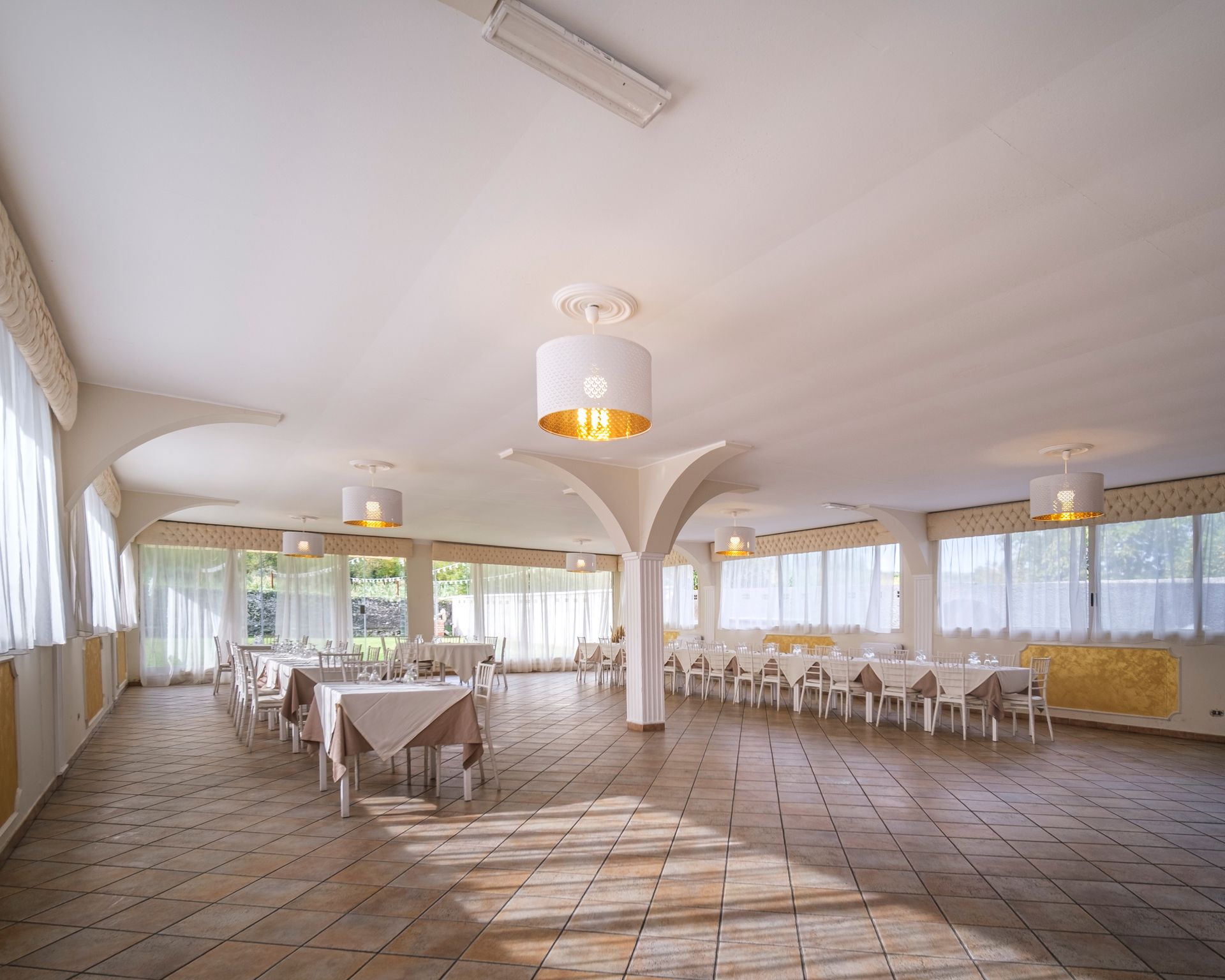 Sala da pranzo vuota e luminosa con tavoli apparecchiati per il pasto; grandi finestre con tende bianche, colori neutri e luci decorative sul soffitto.