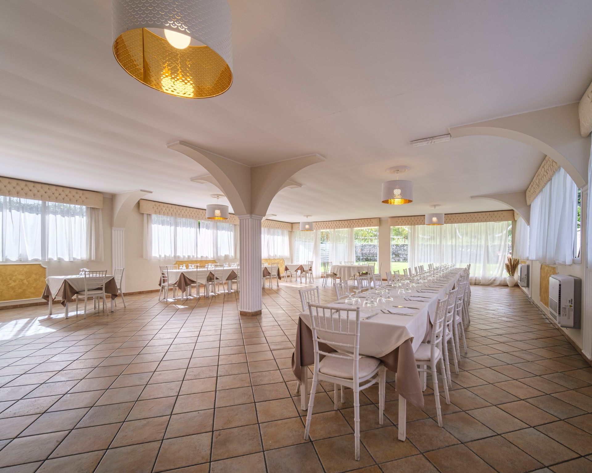 Sala banchetti vuota con lunghi tavoli apparecchiati per il pranzo. Finestre, sedie bianche e pavimento in piastrelle decorate.