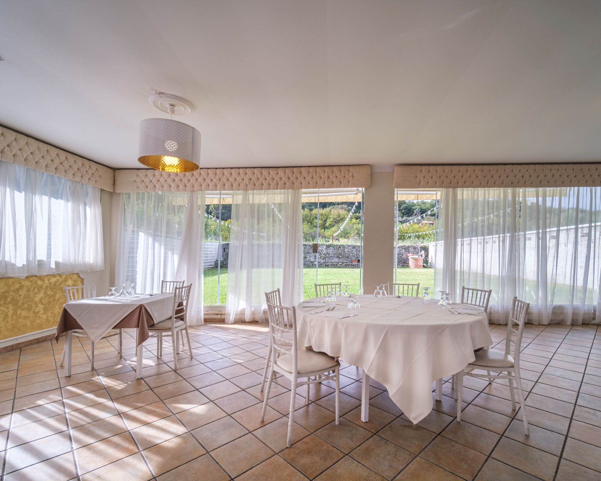 Sala da pranzo con tavoli rotondi e quadrati, tovaglie bianche, sedie e grandi finestre che si affacciano sul prato.