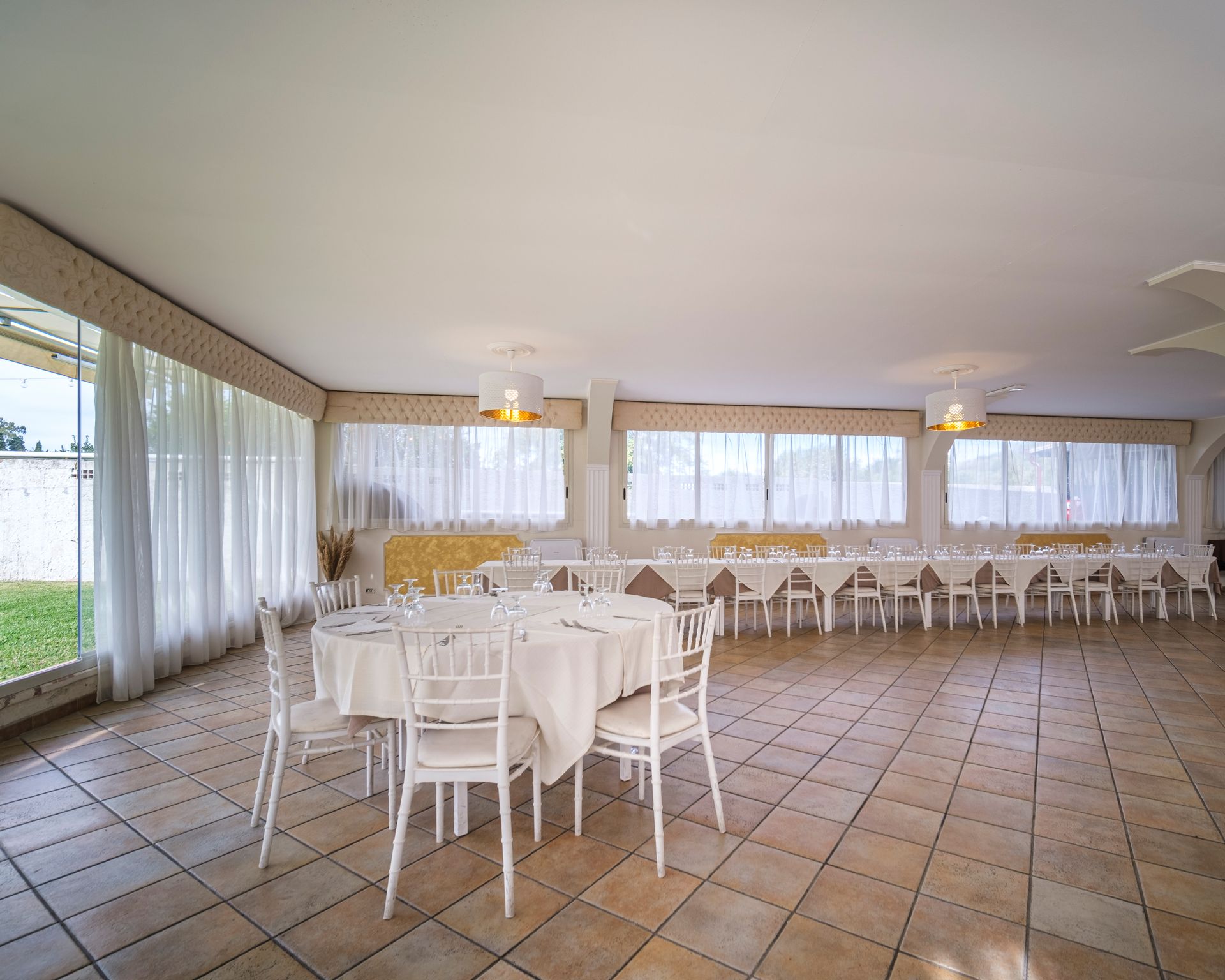 Sala da pranzo vuota con tavoli, sedie e tende bianchi. Pavimento in piastrelle beige e luce naturale intensa.