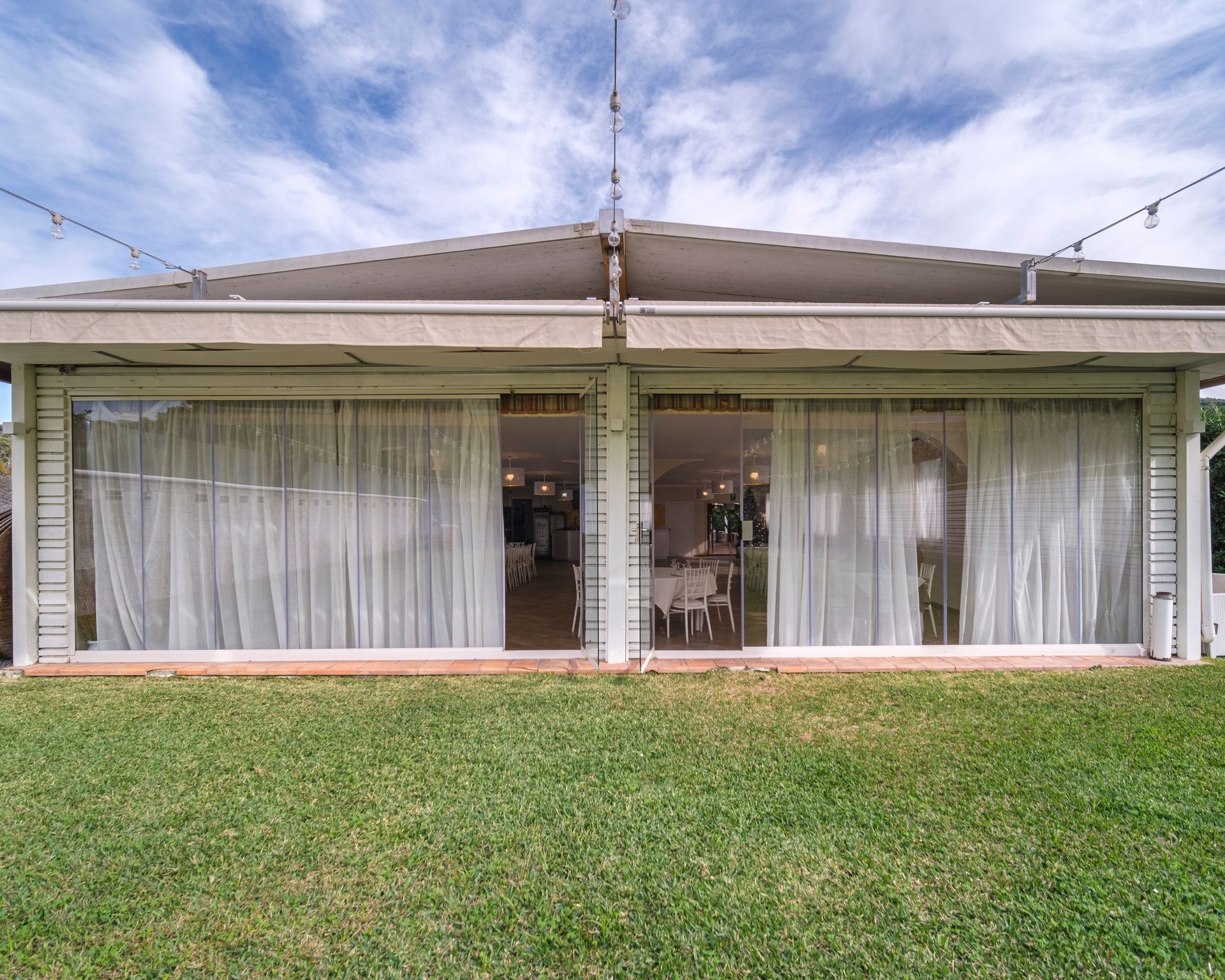 Zona pranzo all'aperto con tende trasparenti, prato verde e cielo azzurro.