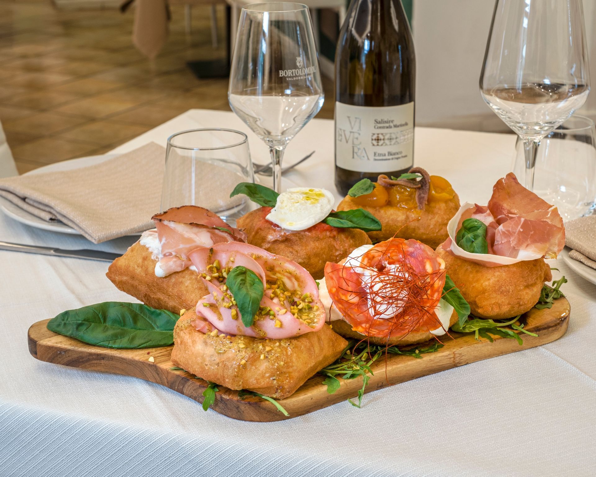 Tagliere di legno con pasta fritta guarnita con vari ingredienti, vino e bicchieri su una tovaglia bianca.