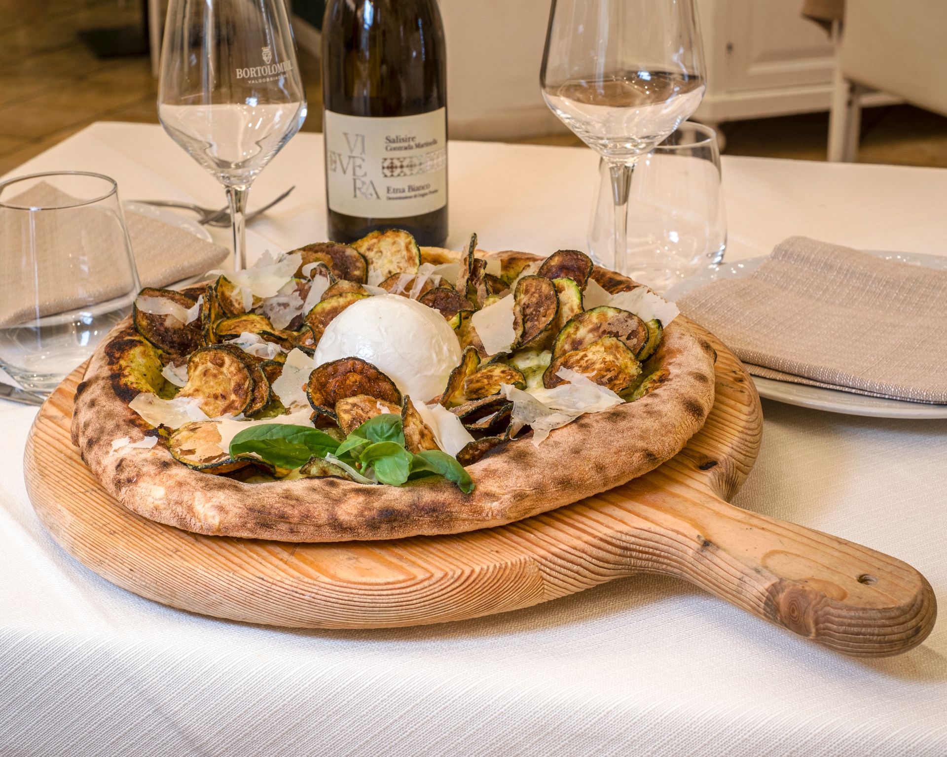 Pizza con condimenti e burrata, su un tagliere di legno con bicchieri da vino e bottiglia, su una tovaglia bianca.