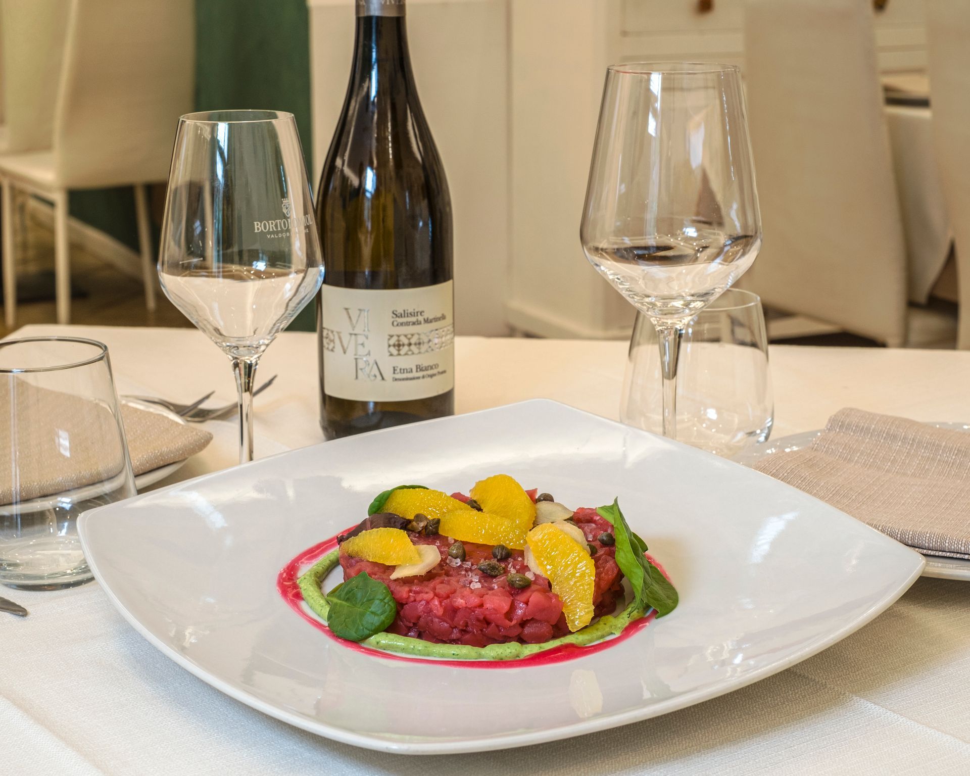 Piatto di cibo con fette d'arancia, bottiglia di vino e bicchieri su una tovaglia bianca.