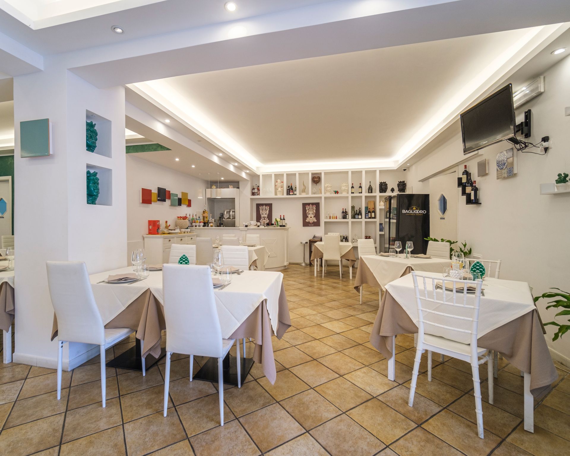 Interno di un ristorante con tavoli bianchi, sedie, piastrelle beige sul pavimento e decorazioni murali.