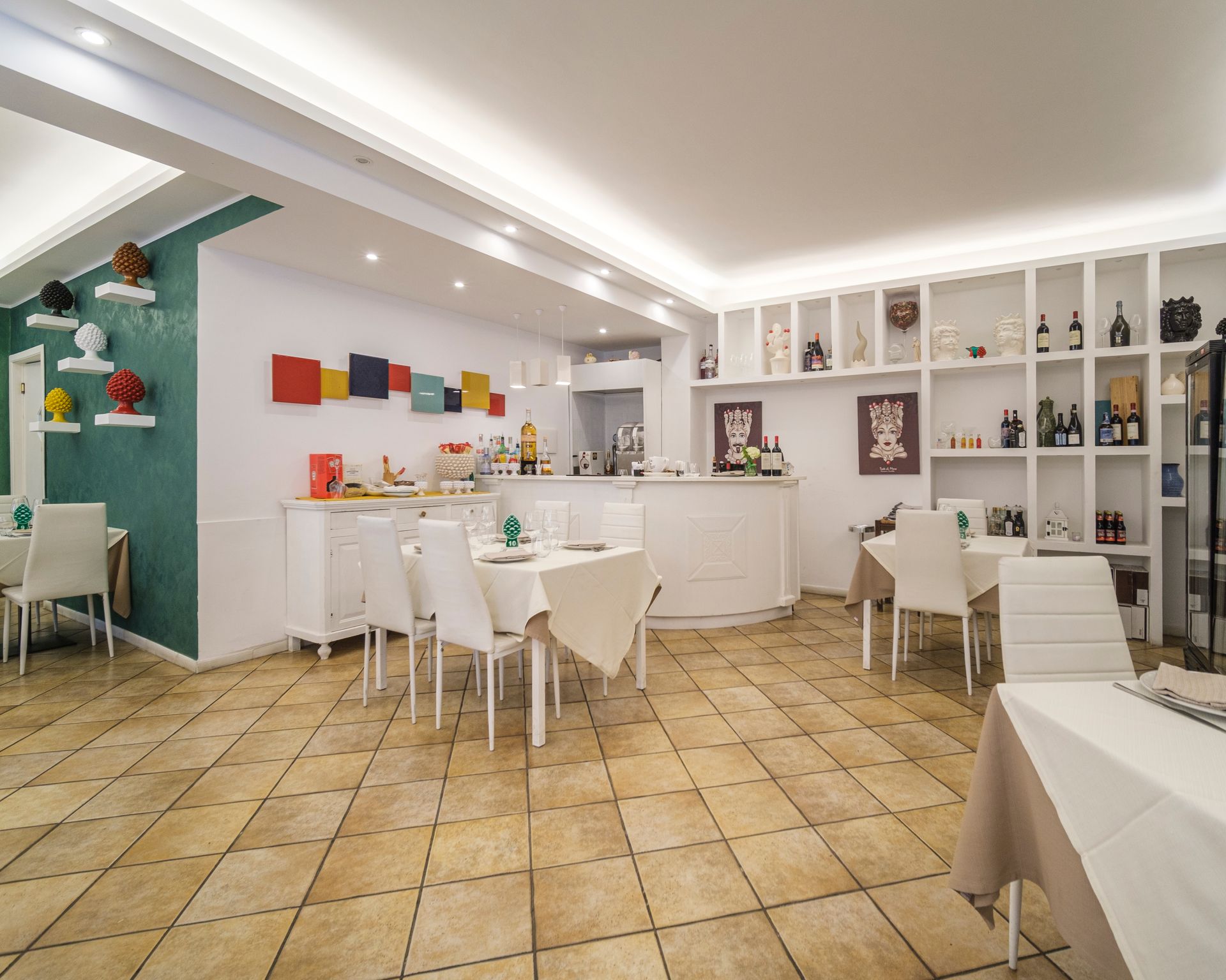 Interno del ristorante con tavoli e sedie bianchi, bar e scaffali decorativi.