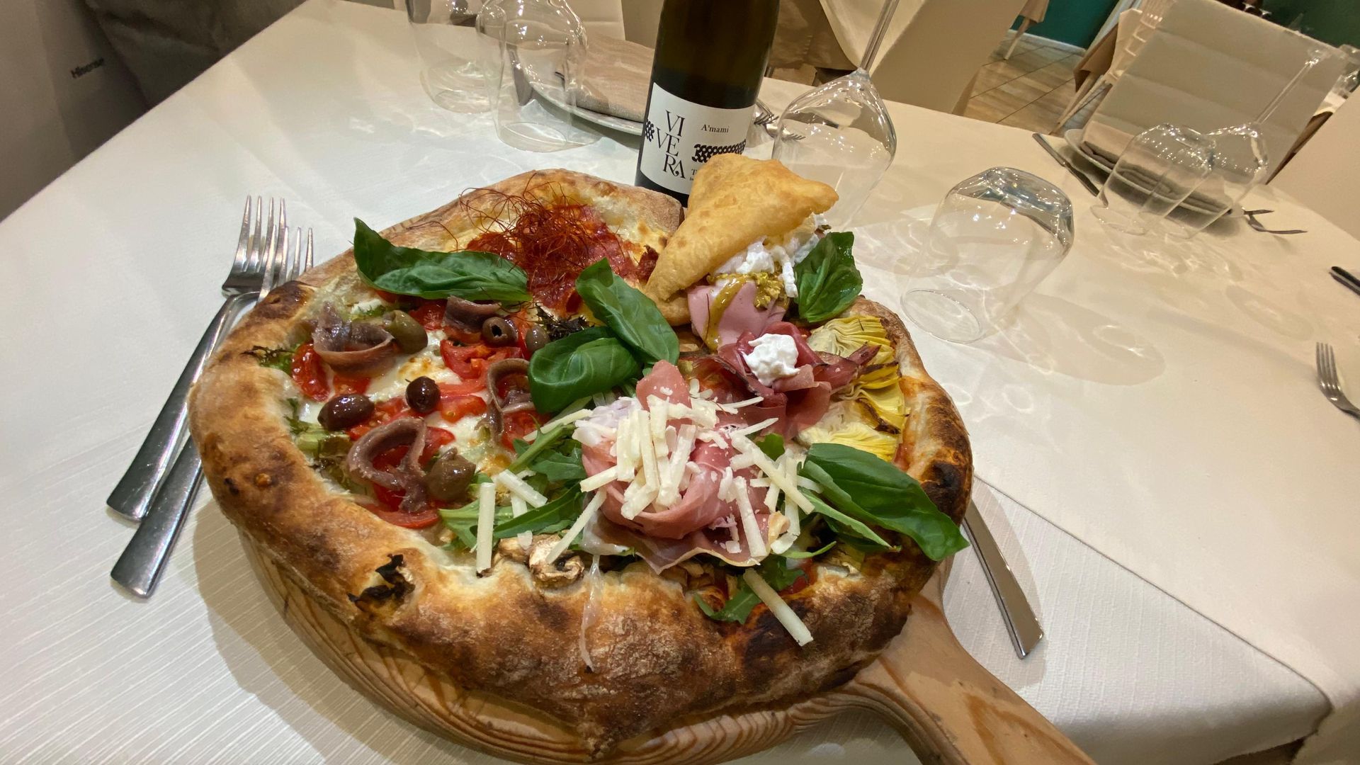Pizza con prosciutto e burrata su un tagliere di legno, con bicchieri di vino e bottiglia.