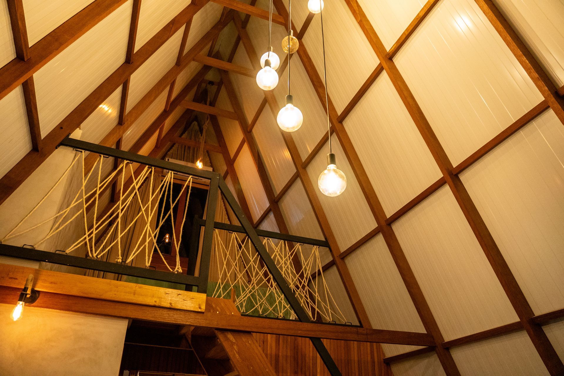 Interior com vigas de madeira, luzes penduradas e um loft com corrimão de corda, vista inclinada. Chalé Mont Hermon.