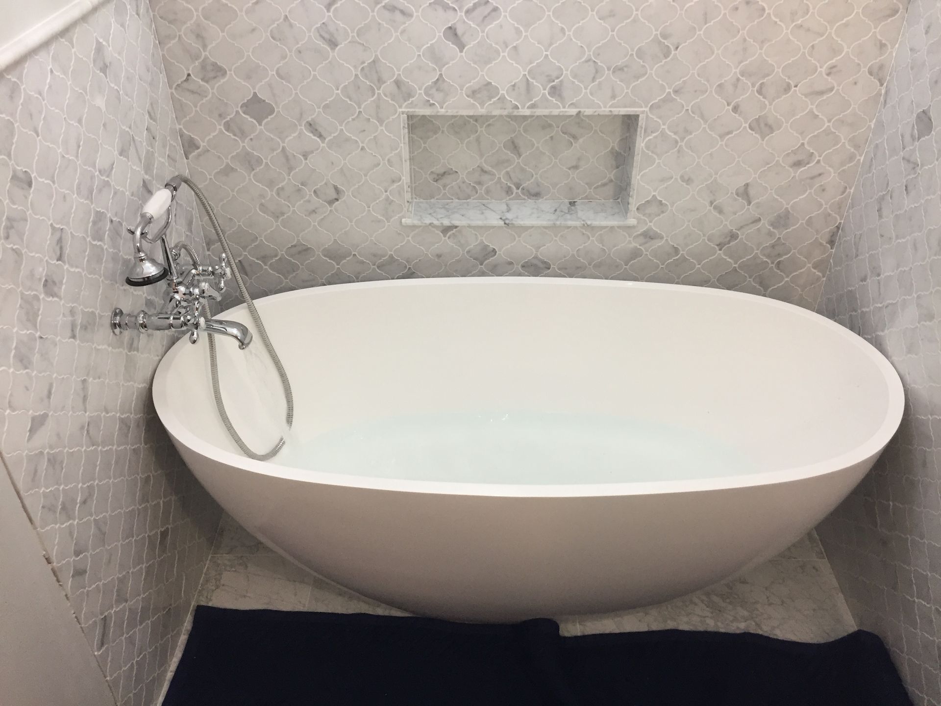 Oval bathtub in a tiled alcove; chrome faucet; gray and white tile; black rug.