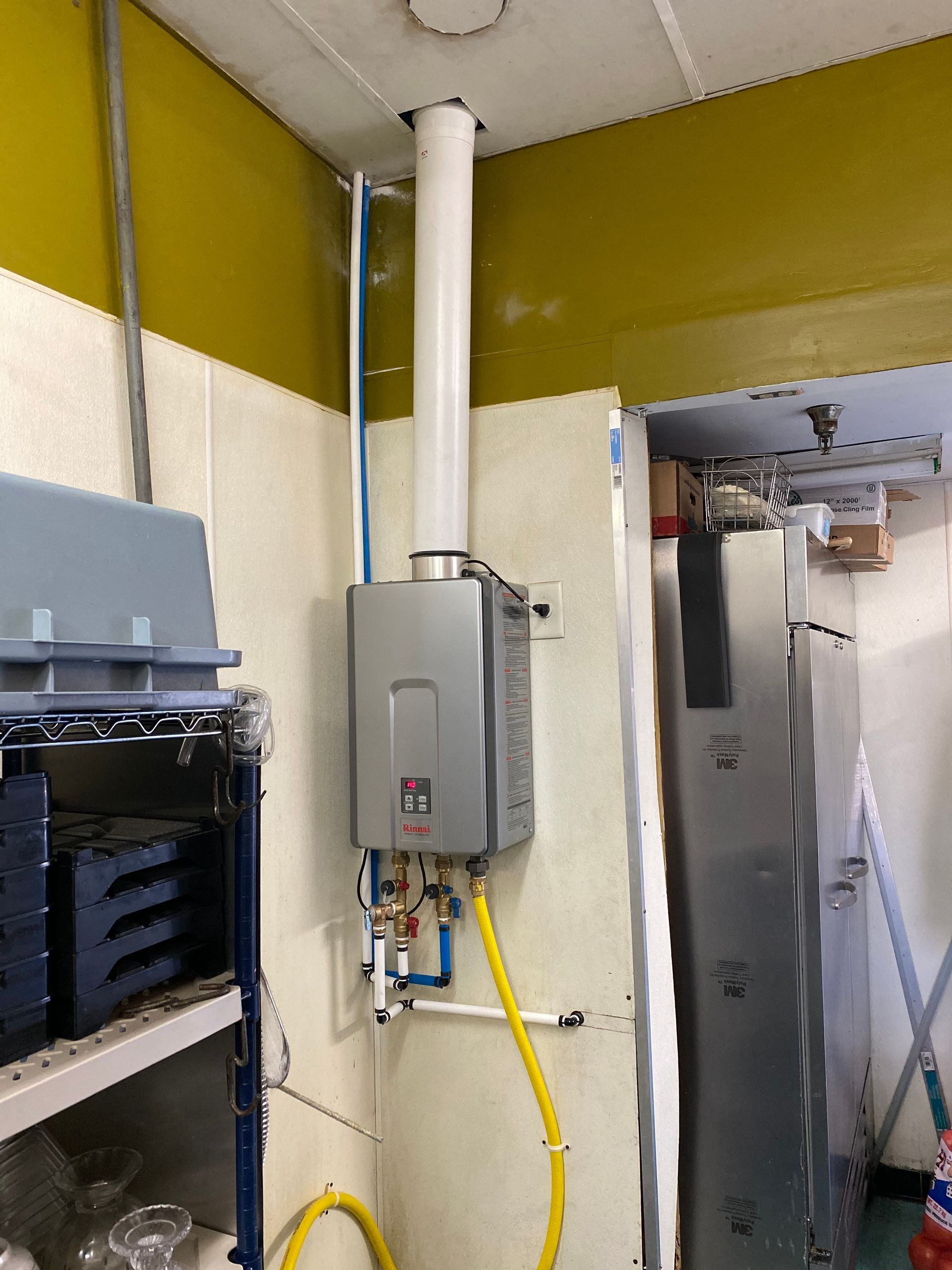 Tankless water heater on a wall, vent pipe to ceiling, blue and yellow pipes, in a utility area.