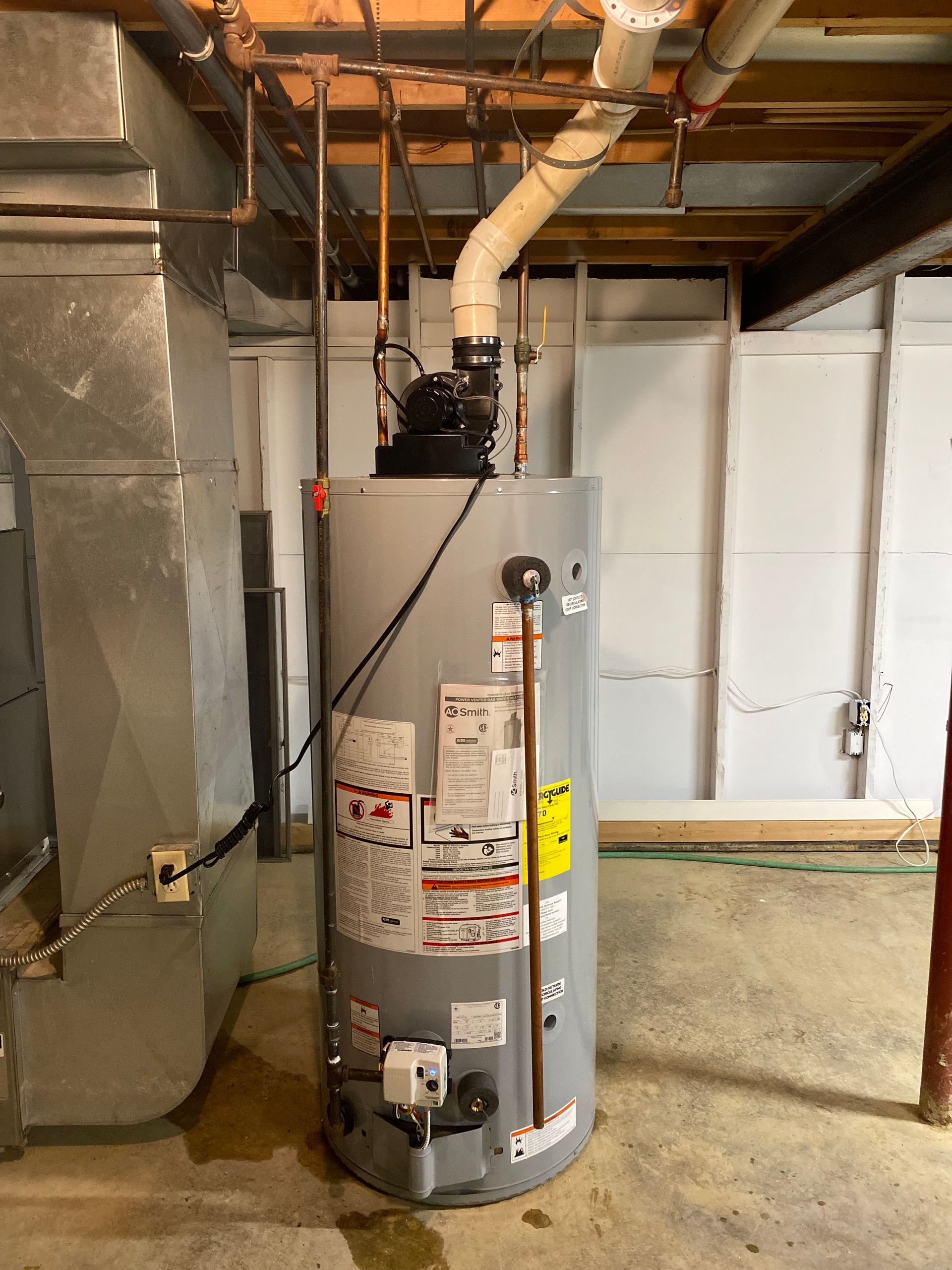A tall, silver water heater in a basement with pipes and ductwork.