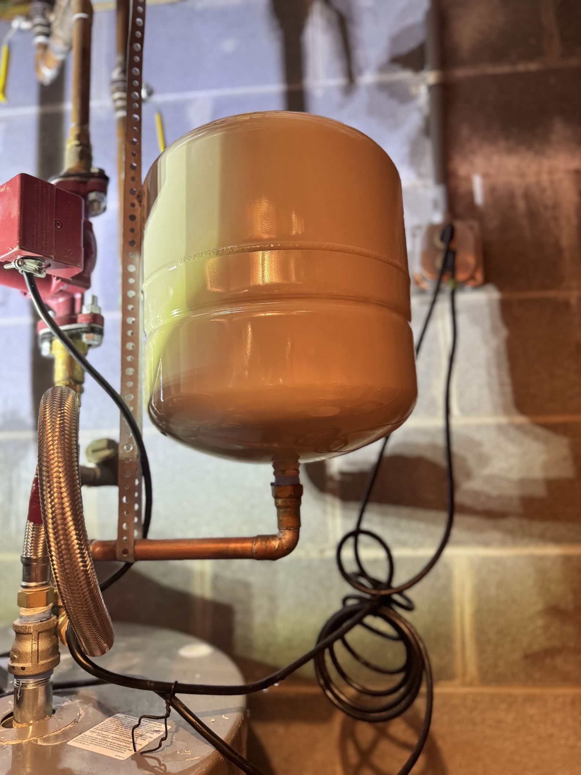 Expansion tank for a boiler system, beige in color, connected by copper pipes and black wiring, indoors.