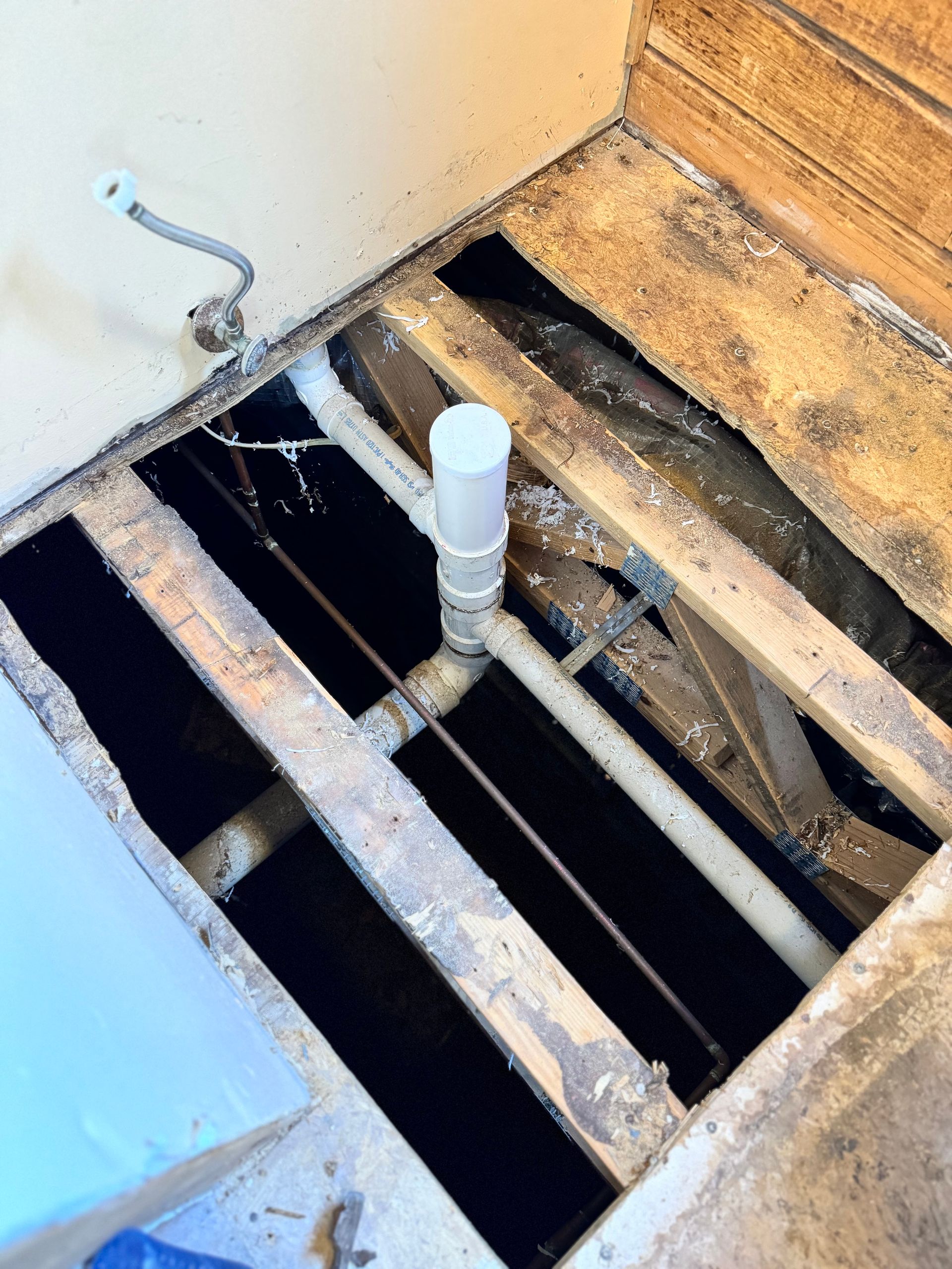 Floorboards removed in bathroom, showing pipes and plumbing. Dark interior with white pipes and wooden supports.