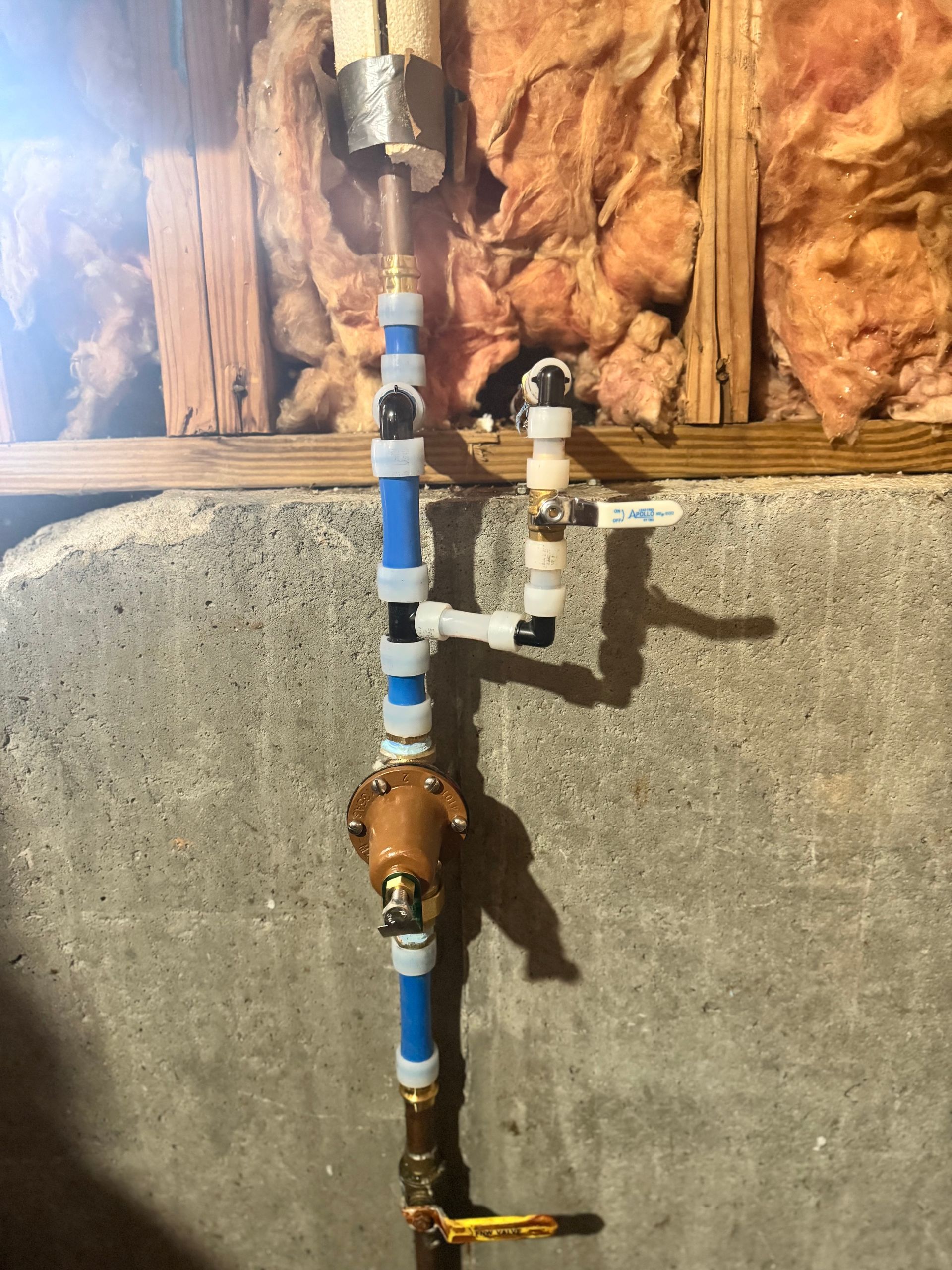 Piping against a concrete wall, with copper and blue PEX tubing, a pressure regulator, and shut-off valves.