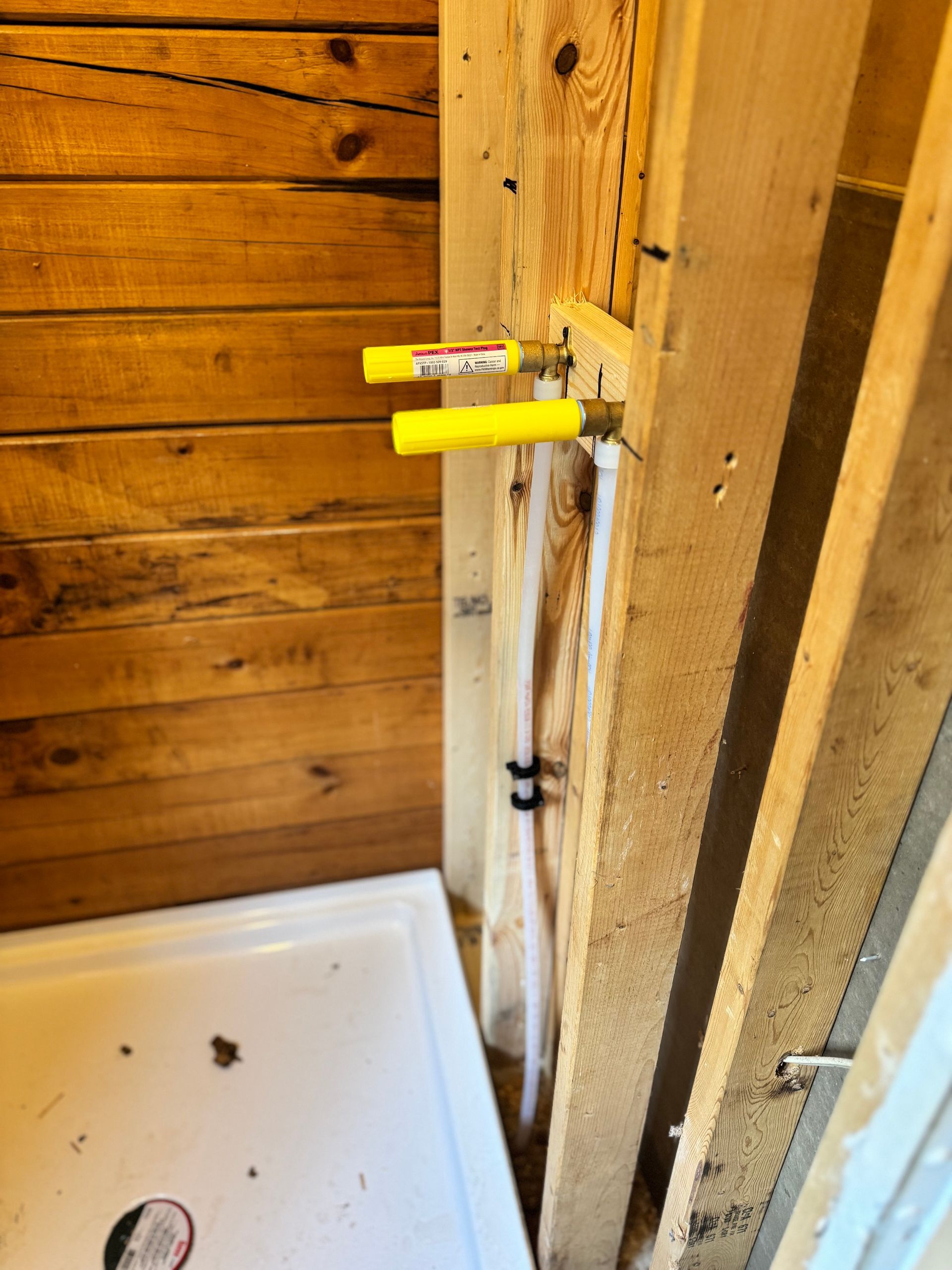 Shower plumbing rough-in with yellow PEX pipes and wood framing, near a white shower base, bathroom construction.