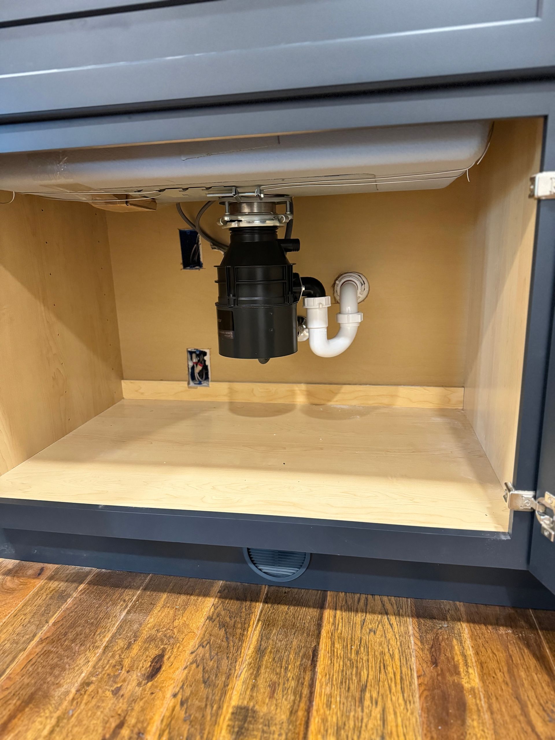 Inside a kitchen cabinet: garbage disposal, sink drain, electrical outlets. Light wood interior, dark blue cabinet exterior.
