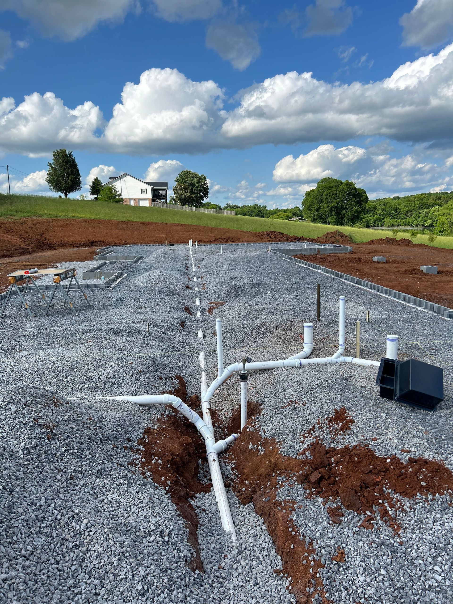 Plumbing pipes laid on gravel at a construction site, with a partially built house in the background under a blue sky.