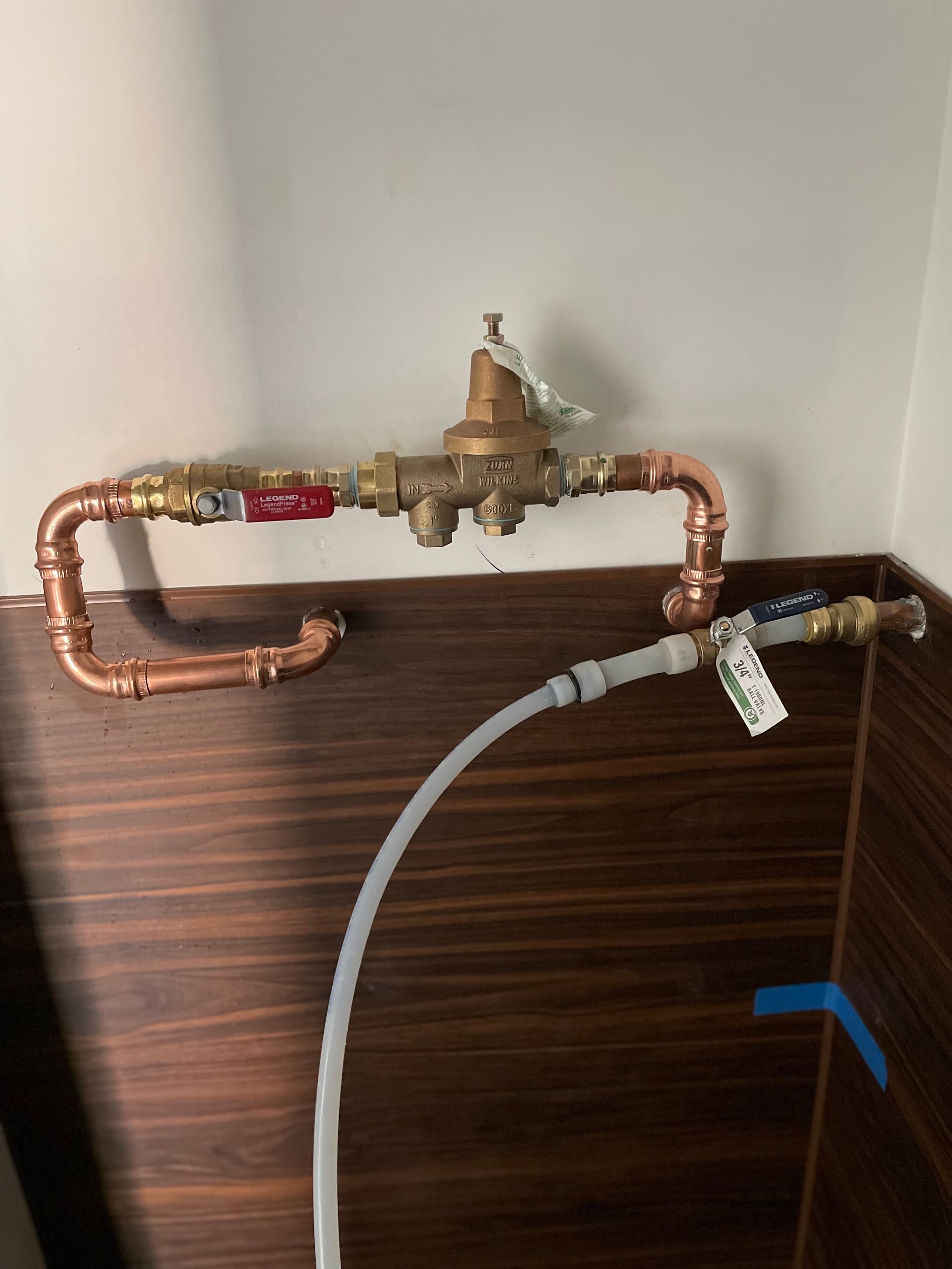 Copper pipes with a pressure-reducing valve, red valve handles, and a clear hose in a wood-paneled space.