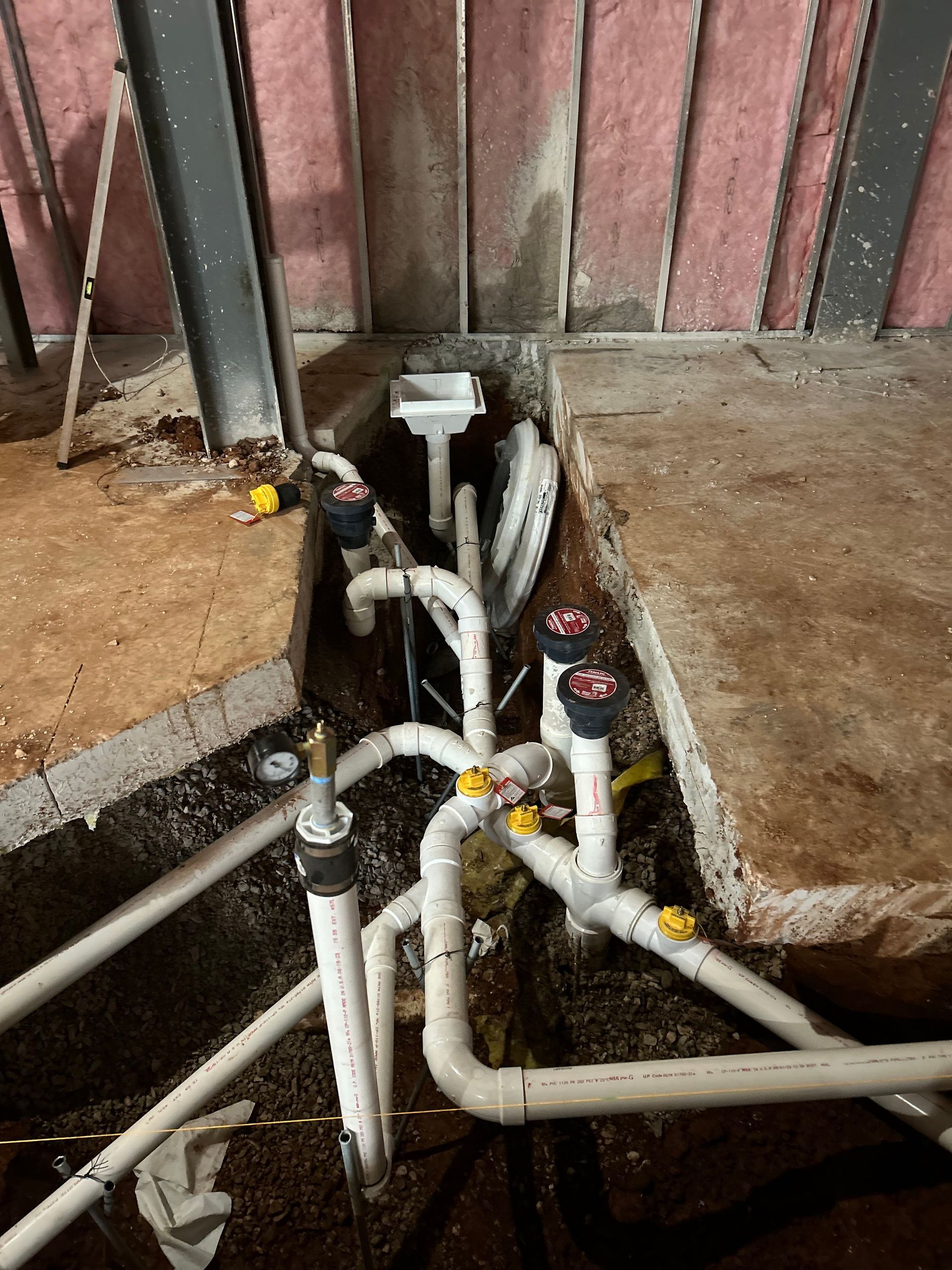 Pipes and plumbing fixtures installed in a trench, with concrete and exposed insulation in a construction setting.
