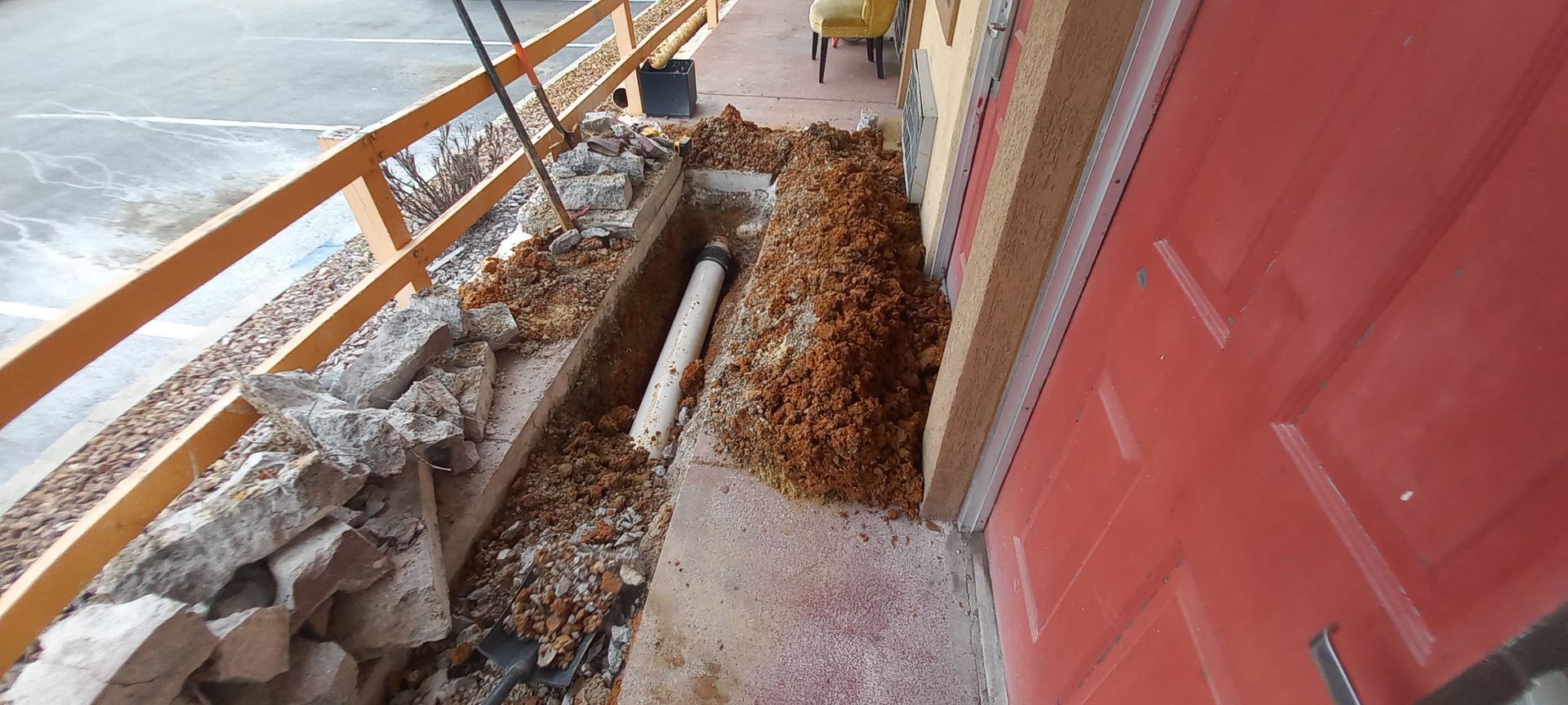 Construction site: Trench next to a red door with debris, exposed pipe, and a wooden railing.