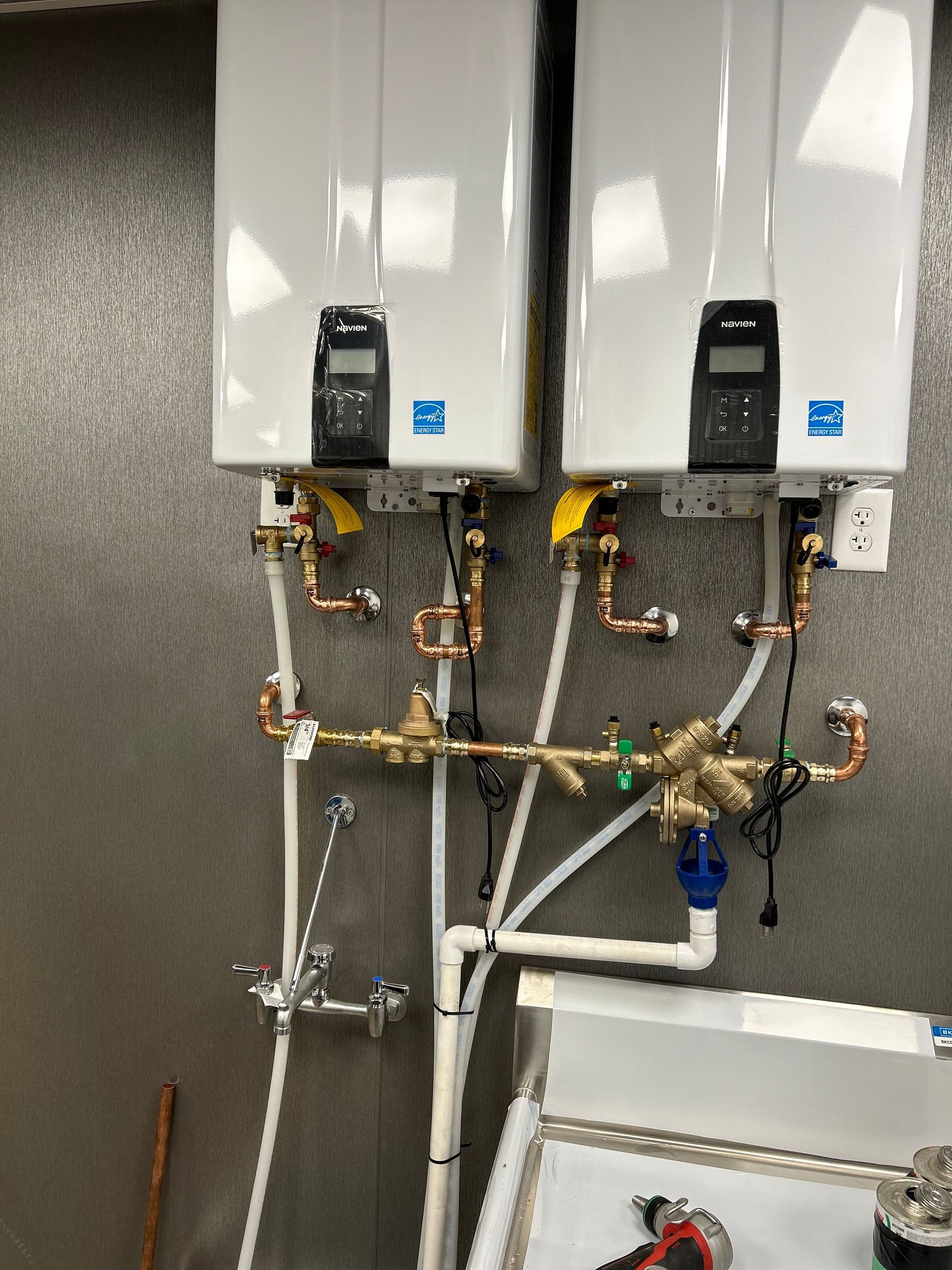 Two white tankless water heaters mounted on a wall with copper and plastic pipes, next to a utility sink.