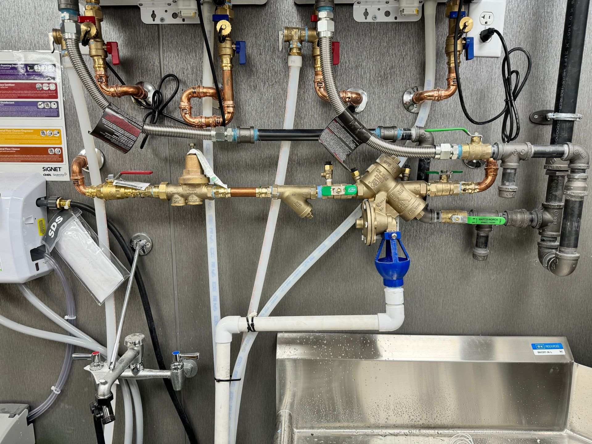 Complex plumbing system with copper and white pipes, valves, and gauges, mounted on a wall above a stainless steel sink.